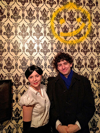 Sherlock and Irene stand against the wall of Sherlock's apartment with the smiley face painted on it.