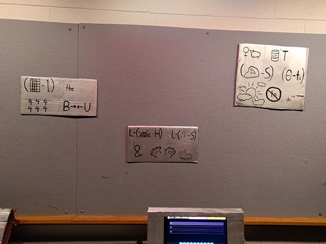 The three password rebus puzzles are mounted above the password entry computer.  The first shows a calendar labeled June, minus one, the word the, a group of fours, and the letters B and U with arrows bringing them together.  The second shows L plus a beehive minus H, L plus music notes minus S, an ampersand, some cat paws, and a cat purring.  The third shows a female symbol next to a sheep, a can with an apostrophe and a letter T, a steak minus S, the greek letter theta minus T A, the sun with clouds and birds flying, the word to inside a no symbol, and the words do and re followed by a blank.
